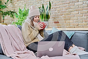 Young woman in sweater hat sitting at home, basking with cup of hot drink