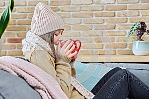 Young woman in sweater hat sitting at home, basking with cup of hot drink