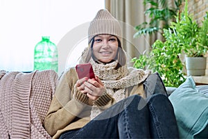 Young woman in sweater hat sitting at home, using smartphone