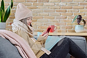 Young woman in sweater hat sitting at home, using smartphone