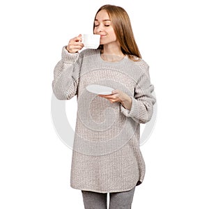 Young woman in sweater holding a white cup and saucer in hands, drinking hot coffee.