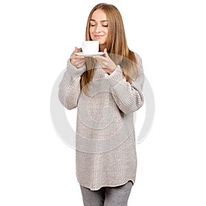 Young woman in sweater holding a white cup and saucer in hands, drinking hot coffee.