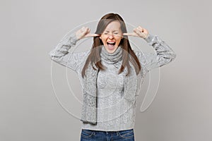 Young woman in sweater, scarf covering ears with fingers, keeping eyes closed, screaming on grey background