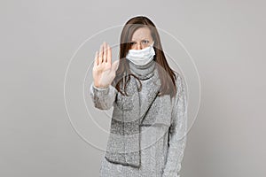 Young woman in sweater, scarf with sterile face mask showing stop gesture with palm isolated on grey background. Healthy