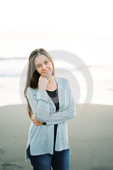 Young woman in a sweater stands on the beach propping her head up with her hand