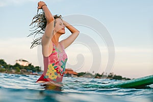 young woman in swimming suit readjusting hair while sitting on surfing board