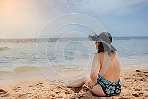 Young woman in swimsuit and a blue big hat sunbathing on the beach