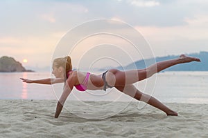 Young woman in swimsuit exercising on beach stretching her legs during sunset at sea. Fitness girl doing exercises on