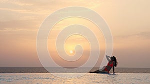 Young woman in swimsuit and sunglasses, is lying and relaxing on edge of outdoor infinity pool with panoramic sea view