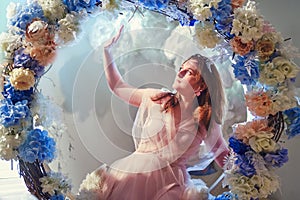 A young woman on a swing of flowers in the rays of light. A woman in a pink dress against a background of mystical luminous clouds