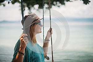 A young woman is swinging on a swing in the shade of a big tree by the sea. Girl on the beach swing on the coast of a tropical