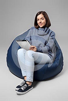 Young woman with tablet PC sitting on beanbag chair against grey background