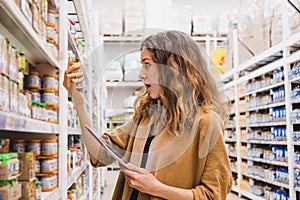 Young woman with a tablet in shock from the composition of baby food in a supermarket, the girl emotionally reads the