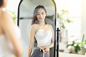 A young woman takes a paper measuring ruler and measures her waist size in the mirror. She is very satisfied with the results of