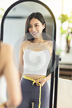 A young woman takes a paper measuring ruler and measures her waist size in the mirror. She is very satisfied with the results of