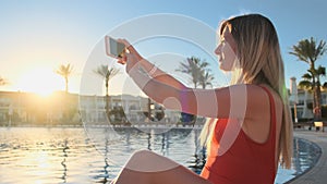 Young woman takes pictures of sunset with phone while relaxing near the swimming pool at luxury villa. Close up of