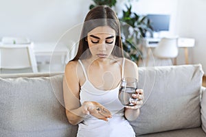 Young woman takes pill with glass of water in hand. Stressed female looking in window and drinking sedated antidepressant meds.