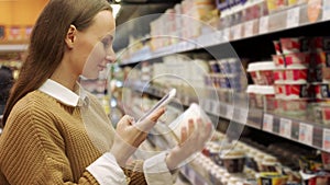 A young woman takes products from a supermarket shelf, holds a smartphone, scans the barcode on the product using a