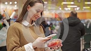 Young woman takes products from the supermarket shelf, holds a smartphone, scans the barcode on the tomato packaging