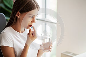 Young woman takes round pill with glass of water in hand