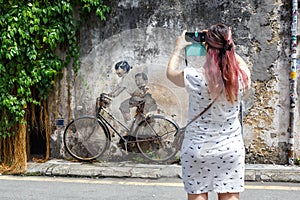 Young woman taking a photo of Street Art mural boy and girl on bicycle on a wall in George Town on Penang island in Malaysia
