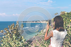 Young woman taking photos on the cliff with a beautiful ocean background at sunny day. Bali island.