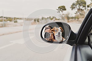 Young woman taking a picture with camera on rear mirror of her cute small jack russell dog watching by the window. Ready to travel