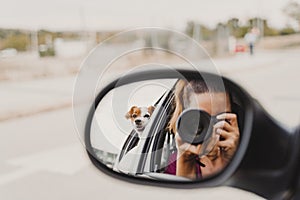 Young woman taking a picture with camera on rear mirror of her cute small jack russell dog watching by the window. Ready to travel