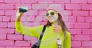 Young woman taking selfie picture by phone with lollipop on a pink brick wall background