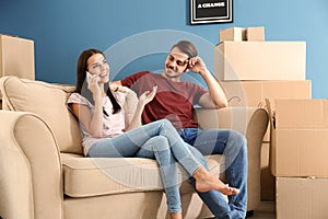 Young woman talking by mobile phone while her husband sitting on sofa at new home