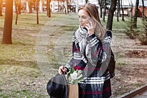 Young woman talking with parents on the phone walking on a sunny day in the park. Beautiful girl in a coat, hat and
