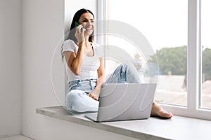 Young woman talking phone and sitting on windowsill at home