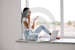 Young woman talking phone and sitting on windowsill at home