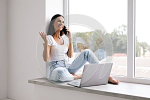 Young woman talking phone and sitting on windowsill at home