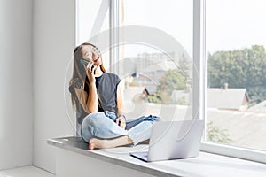 Young woman talking phone and sitting on windowsill at home