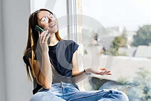 Young woman talking phone and sitting on windowsill at home