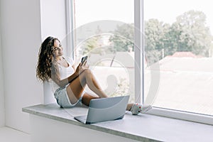 Young woman talking phone and sitting on windowsill with laptop computer at home