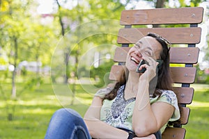 Young woman talking on the smartphone and laughing in the park on the bench Beautiful female relaxing on a park bench and using a