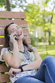 Young woman talking on the smartphone and laughing in the park on the bench Beautiful female relaxing on a park bench and using a