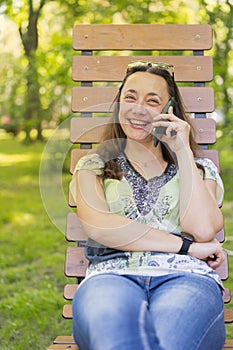 Young woman talking on the smartphone and laughing in the park on the bench Beautiful female relaxing on a park bench