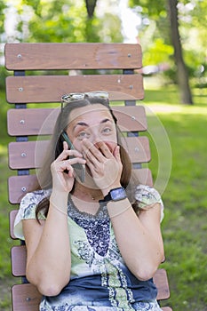 Young woman talking on the smartphone and laughing in the park on the bench Beautiful female relaxing on a park bench and using a