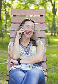 Young woman talking on the smartphone and laughing in the park on the bench Beautiful female relaxing on a park bench and using a