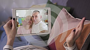Young woman talking to her smiling parents facetime with tablet while lying on sofa
