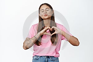 Young woman with tattoos pucker lips for kiss, shows heart hand sign and close eyes, standing silly against white