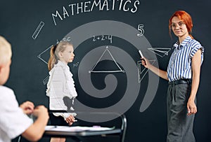 Young woman, teacher of math standing by blackboard with little girl student and solving geometry tasks. Lesson