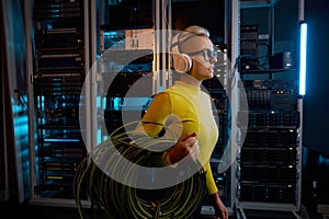 Young woman IT technician walking with wires through server rack in data center