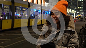 Young woman teenager using her cell phone at night with trams by Alexanderplatz Station, Berlin, Germany