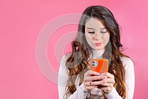 Young woman texting someone unhappily using smartphone on the pink background