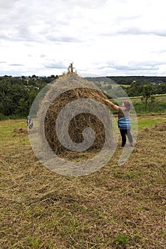 A young woman throws hay on a wooden support with a hand tool for further drying on the field, the season of haymaking