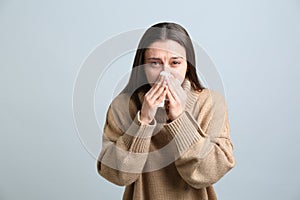 Young woman with tissue suffering from runny nose on light grey background